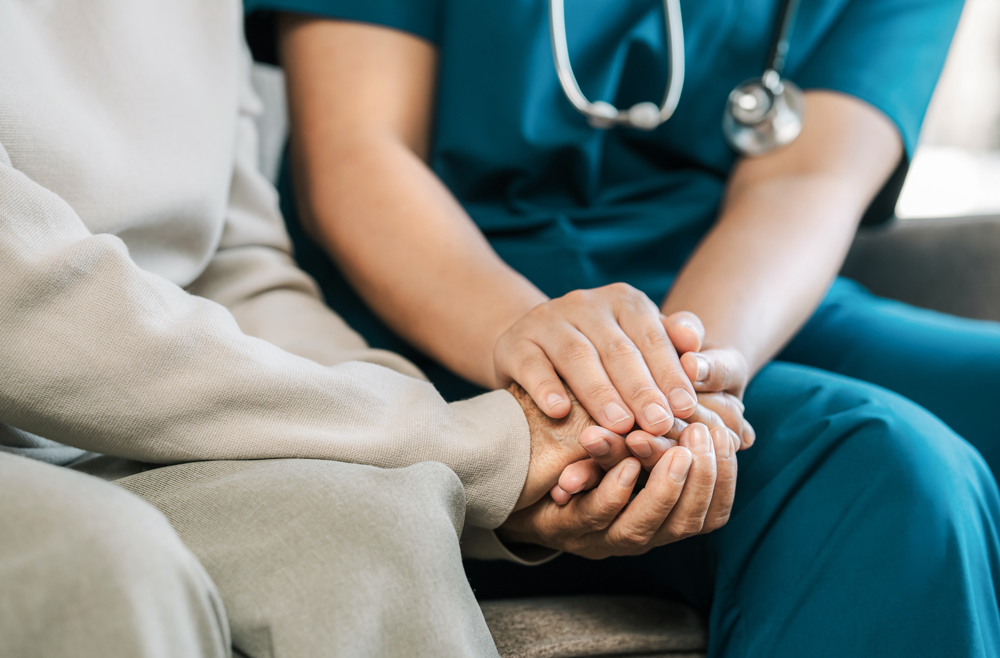 A nurse sits next to a patient on a couch, holding their hands.